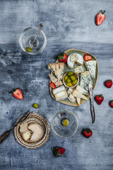 variety of cheese with fresh strawberries, grape, olives and nuts, martini glasses on shabby blue background, top view, party appetizer concept