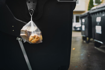 A wet plastic bag with old pastries hanging on the side of the waste container.
