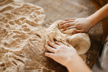 Child making dough for bread, punching it and shaping into a ball, on a wooden table surface.