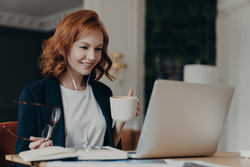 Young female teacher conducts lesson online, talks wih pupils via laptop computer, organizes video conference, teaches students, drinks aromatic coffee, holds glasses in hand, dressed formally