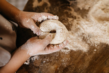 Child making dough for bread, shaping into a bagel on a wooden table surface.