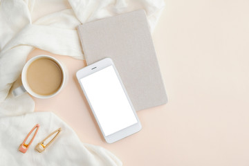 Smartphone mockup with blank screen, cup of coffee, paper notebook, blanket and feminine accessories on beige background. Home office desk table concept. Flat lay, top view.