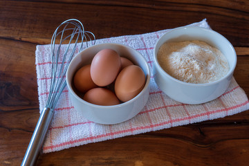 Raw chicken eggs on white bowl with flour and with whisk on wooden background. Ready to start the cooking concept - Powered by Adobe