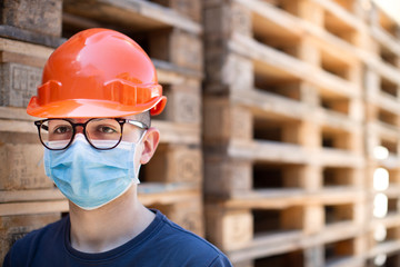 Giovane operaio con casco protettivo arancione, mascherina chirurgica  e pallet accatastati di fianco, guarda deciso