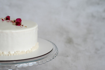 Close up view of simple white mousse cake decorated with dried rose buds  on smooth glaze. Сake stands on a round transparent glass stand on a gray texture background with place for text.