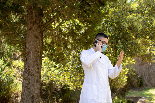 Giovane medico con camice bianco e mascherina protettiva parla al cellulare isolato su uno sfondo di un bosco 