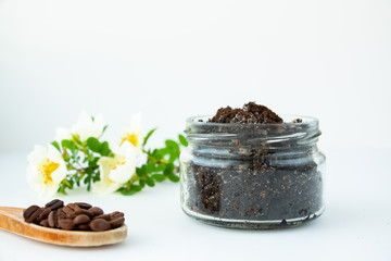 Coffee and sugar scrub in a round glass jar. Wooden spoon with coffee beans and a flowering branch on a white background. The concept of home spa care and beauty.