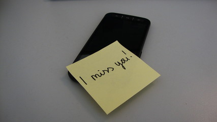 A handwritten note reading "I miss you!" sticking to a black blank mobile phone on a white background 