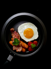 Pan of fried eggs,bacon,sausage and cherry-tomatoes on black background,top view
