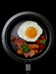Pan of fried eggs,bacon,sausage and cherry-tomatoes on black background,top view