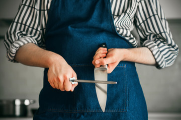 The cook, dressed in a blue apron and striped shirt, is standing in the kitchen sharpening a knife blade with a whetstone.