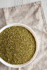Dried Green Greek Oregano Spice in a white bowl on cloth, view from above. Flat lay, top view, overhead. Copy space.