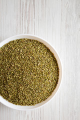 Dried Green Greek Oregano Spice in a white bowl on a white wooden background, top view. Flat lay, overhead, from above. Copy space.