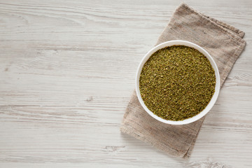 Dried Green Greek Oregano Spice in a white bowl on a white wooden surface, top view. Flat lay, overhead, from above. Copy space.