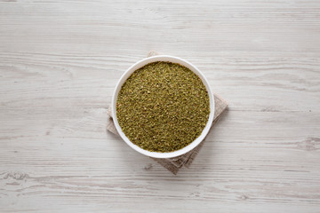 Dried Green Greek Oregano Spice in a white bowl on a white wooden background, top view. Flat lay, overhead, from above.