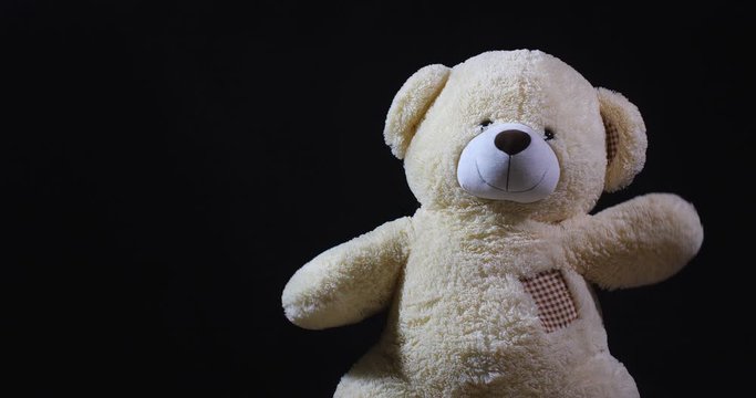 A lively toy bear looking at the camera and waving its paws as a sign of welcome on a black studio background with dramatic light and free space for inscription.