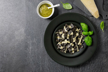 Italian farfalle black pasta, on a dark background stone, black plate. Ingredients for cooking. Hard Parmesan, Basil, Pesto. Top view. Copy space.
