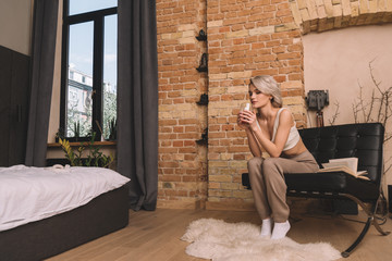 pensive, beautiful woman holding glass of wine while sitting in armchair in bedroom