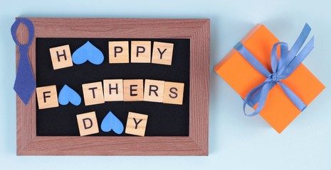 Father's Day greeting card message with paper tie, beautiful gift box and frame on blue desk.Flat lay composition with phrase HAPPY FATHER'S DAY.Party decoration concept.