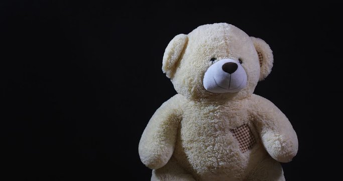 A lively toy bear looking at the camera and twisting his head in denial as if he is saying no on a black studio background with dramatic light and free space for inscription.
