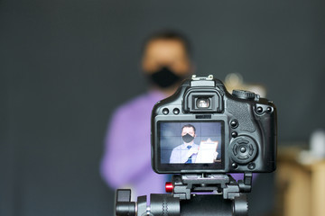 A blogger in a protective mask takes pictures of himself on camera. Advertises a product. Focus is on his camera on a tripod and the image on it. Everything else is blurry.