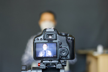 A blogger in a protective mask takes pictures of himself on camera. Focus is on his camera on a tripod and the image on it. Everything else is blurry.