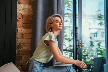 dreamy, beautiful woman looking away while sitting near window with laptop
