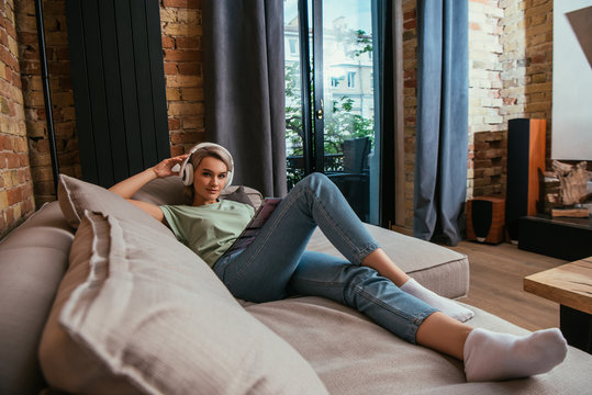 selective focus of beautiful woman in wireless headphones looking at camera while chilling on sofa