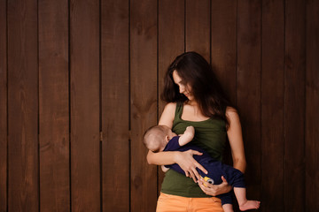 Fototapeta premium Young woman holding a newborn baby in her arms on the brown background. Newborn photosession. Family portrait. Mother is kissing her baby. Baby goods packing template. Nursery. Medical and healthy.