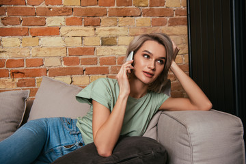 Fototapeta na wymiar beautiful woman looking away while sitting on sofa and talking on smartphone