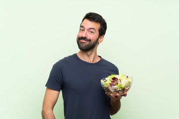 Young handsome man with salad over isolated green wall laughing