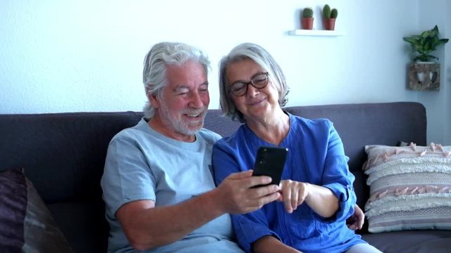two seniors or pensioners at home on the sofa having fun together looking some funny videos or photos on the phone - laughing and having fun together
