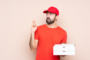 Young man holding a pizza over isolated background with fingers crossing and wishing the best