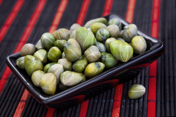 Capers in a black plate on a tablecloth with red stripes. Bunch canned pickled or salted capers.