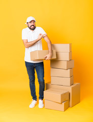 Full-length shot of delivery man among boxes over isolated yellow background frightened and pointing to the side