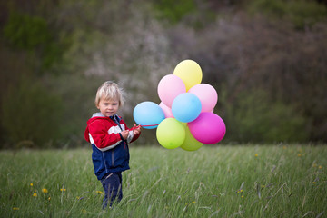 Little child, cute boy on a spring cold windy rainy day, holding colorful balloons in a field, running