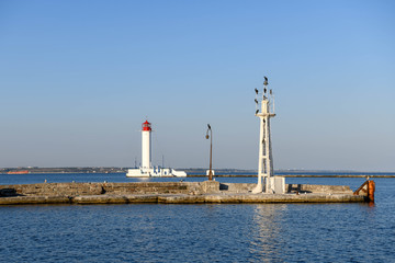 Birds are perched on a tower and the Odessa lighthouse is in the background illuminated by the setting sun.