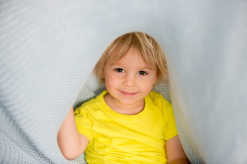 Naklejka premium Little toddler boy with yellow shirt, hiding under blanket, smiling