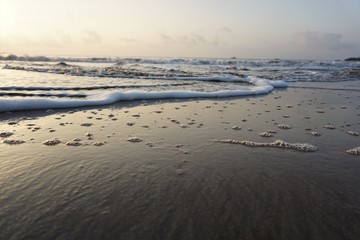 Rolling waves on the seashore with froth and bubbles on the wet sand during dusk or dawn taken from a low angle with scattered clouds over horizon.