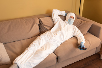 Man in medical mask, hazmat suit and latex gloves watching tv on sofa at home