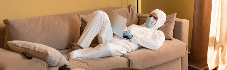 KYIV, UKRAINE - APRIL 24, 2020: Panoramic shot of man in hazmat suit and medical mask using laptop near remote controller and joystick on sofa