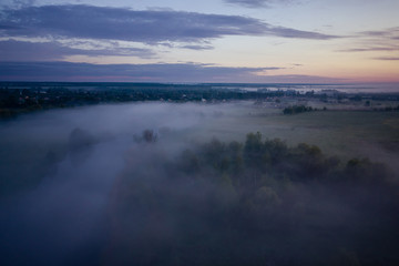 The river and meadow are covered with fog until sunrise. Pre-dawn, hazy landscape. Trees peek through the fog.