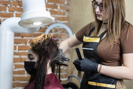 A girl hairdresser makes a client keratin hair straightening using a straightener under the hood from formaldehyde fumes. The girl is sitting in a mask in the salon under the keratin hood.