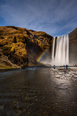 Skogafoss waterfall in South Iceland with a beautiful rainbow. Tourists admiring the waterfall. Popular and famous tourist attraction.