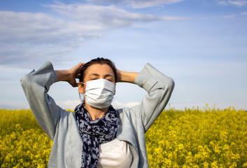Fototapeta premium Woman in a medical mask on a background of a yellow flower field. COVID-19 Pandemic Coronavirus concept. Sky with clouds and sun.