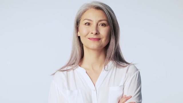An attractive old mature woman with long gray hair wearing formal business clothes is looking to the camera standing isolated over white background in studio