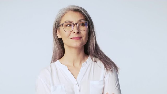 A beautiful old mature woman with long gray hair wearing glasses is looking to the side while thinking about something standing isolated over white background in studio