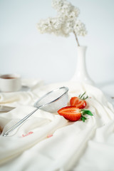 Sliced strawberry, lying next to a sieve for powdered sugar, on a white cloth, near the flowers of lilac. Food photography. Advertising and commercial close up design.