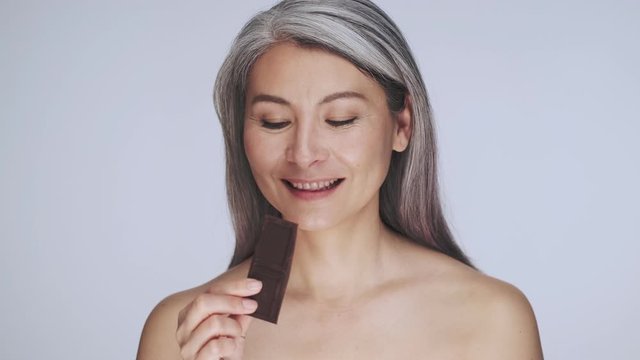 A Happy Old Mature Half-naked Woman With Long Gray Hair Is Eating Eat A Bar Of Chocolate Isolated Over White Background In Studio