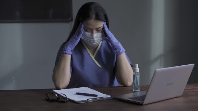 A person sitting at a desk in front of a laptop with a tired face and holding head with cleaned antibacterial liquid gloves. Headache at work nurse pains. Prores 422.